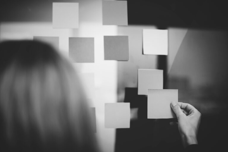 Photo en noir et blanc d’une personne organisant des notes autocollantes sur une paroi en verre, illustrant la planification, l’organisation et la concentration.
